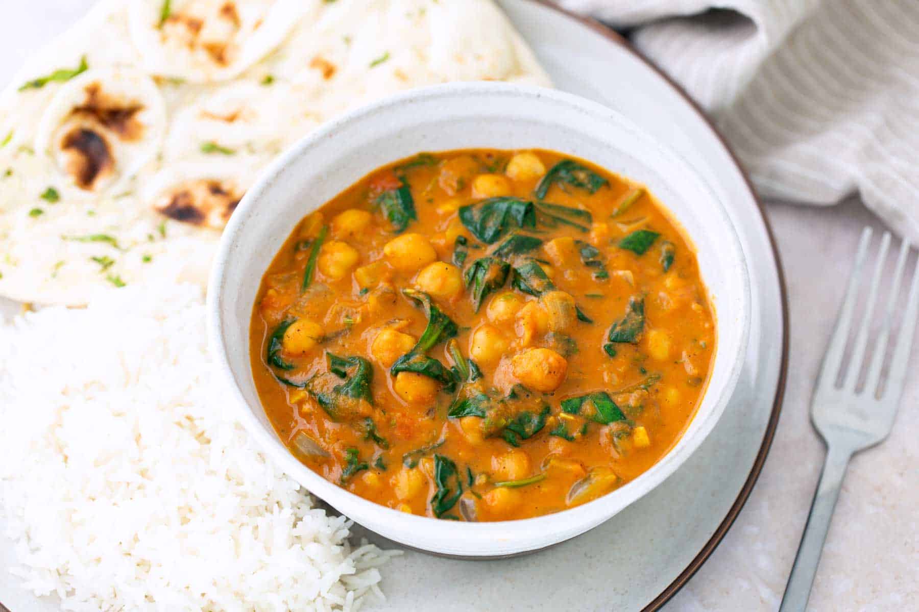 A bowl of chickpea and spinach curry with white rice and naan bread on a plate, next to a fork and a napkin.
