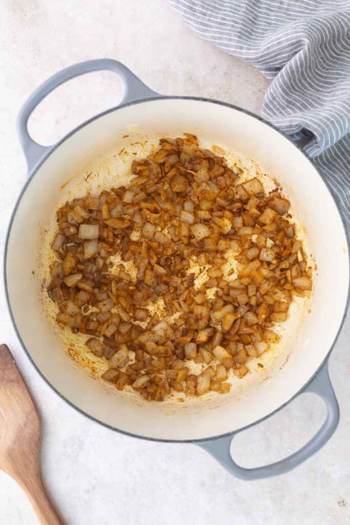 Chopped onions and spices are sautéing in a large white pot, with a wooden spoon and a striped towel nearby on a light surface.