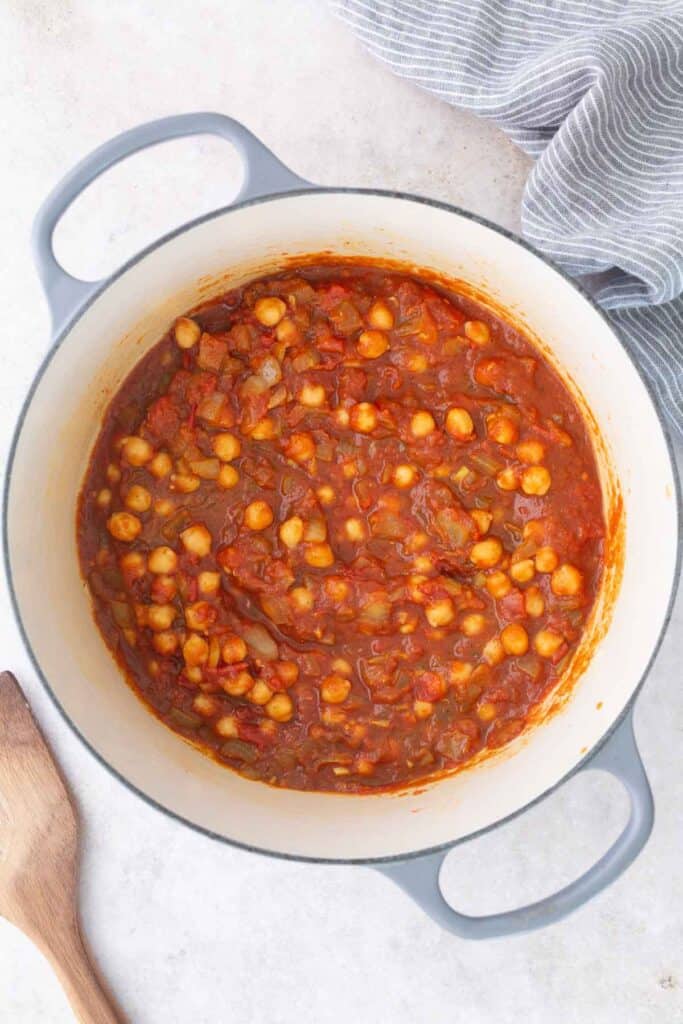 A pot filled with a tomato-based chickpea stew sits on a light countertop, with a wooden spoon and striped cloth beside it.