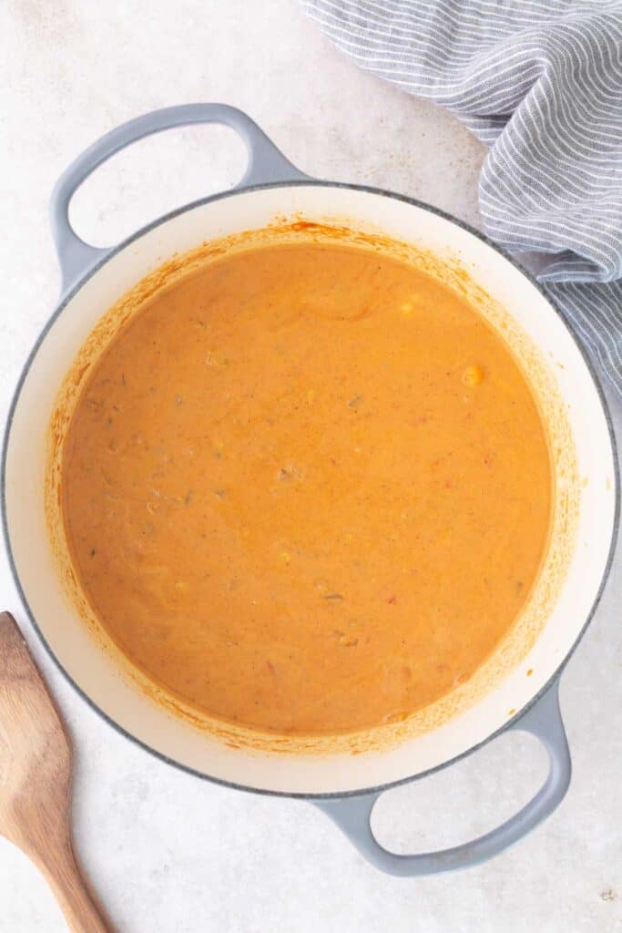 A pot of orange-colored soup or sauce sits on a light surface next to a wooden spoon and a gray-striped cloth.