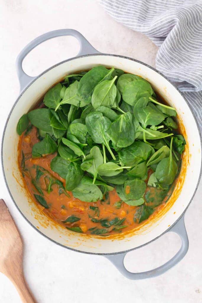 A white pot containing a creamy orange stew or curry with fresh spinach leaves on top, next to a wooden spoon and a striped cloth.