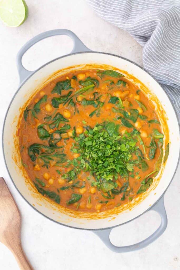 A pot of chickpea and spinach curry in a creamy orange sauce, topped with chopped cilantro. A wooden spoon, a lime half, and a striped cloth are nearby.