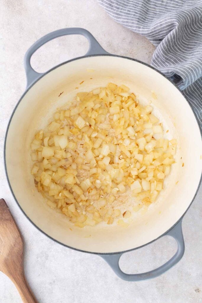 Diced onions being sautéed in a large light-colored pot, with a wooden spoon and striped cloth nearby on a light countertop.