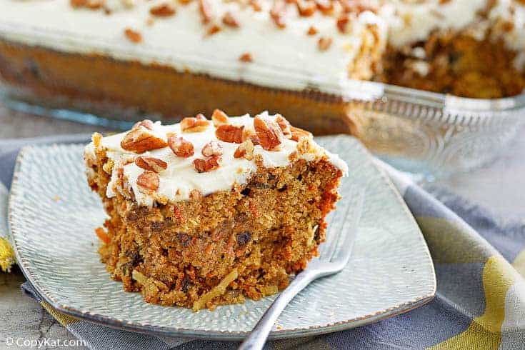 A slice of carrot cake with cream cheese frosting and chopped pecans on top sits on a plate with a fork; the rest of the cake is visible in a glass pan in the background.