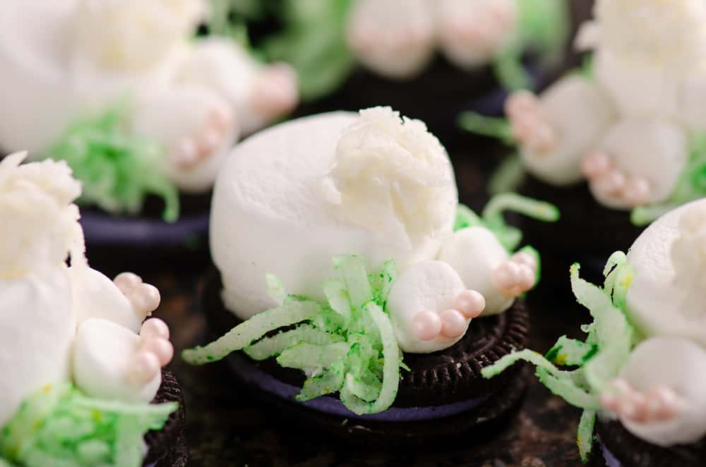 Cookies decorated to look like bunny butts with marshmallows, pink candy, and shredded green coconut, arranged on a dark surface.