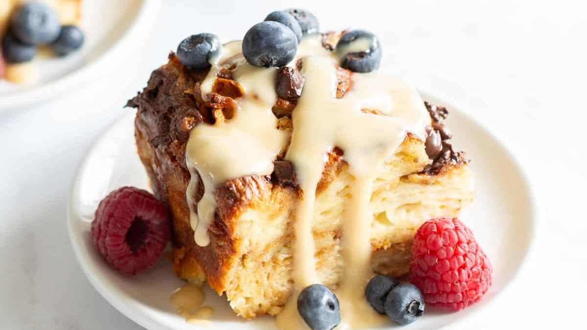 A plate of croissant bread pudding topped with cream and blueberries, served with raspberries, beside a blueberry basket and a croissant.