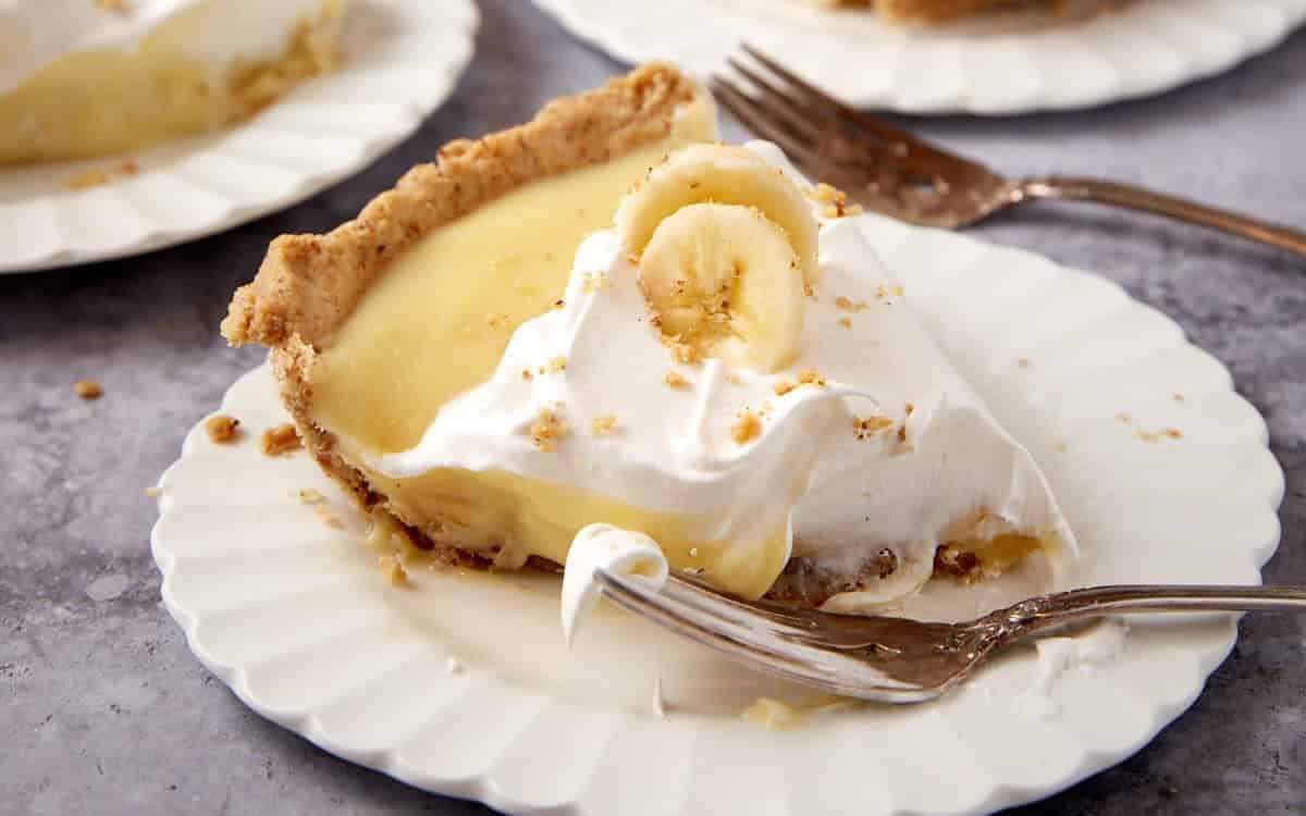 A slice of banana cream pie topped with whipped cream and a banana slice, served on a white plate with a fork.