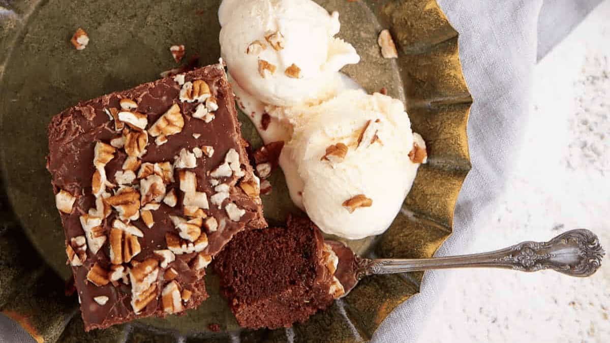 A slice of chocolate cake topped with chopped nuts is served on a plate with two scoops of vanilla ice cream and a spoon.