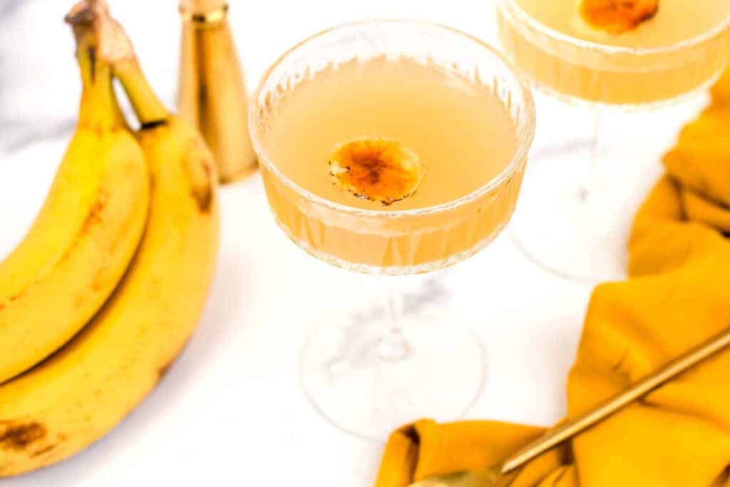 A cocktail in a coupe glass garnished with a banana slice, with bananas, a gold spoon, and a gold shaker on a white surface.
