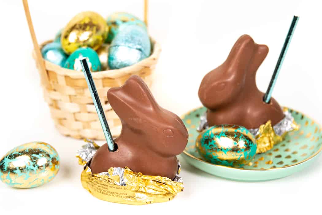 Two chocolate bunny pops with straws are placed on a plate and next to foil-wrapped chocolate eggs, with a basket of decorated eggs in the background.