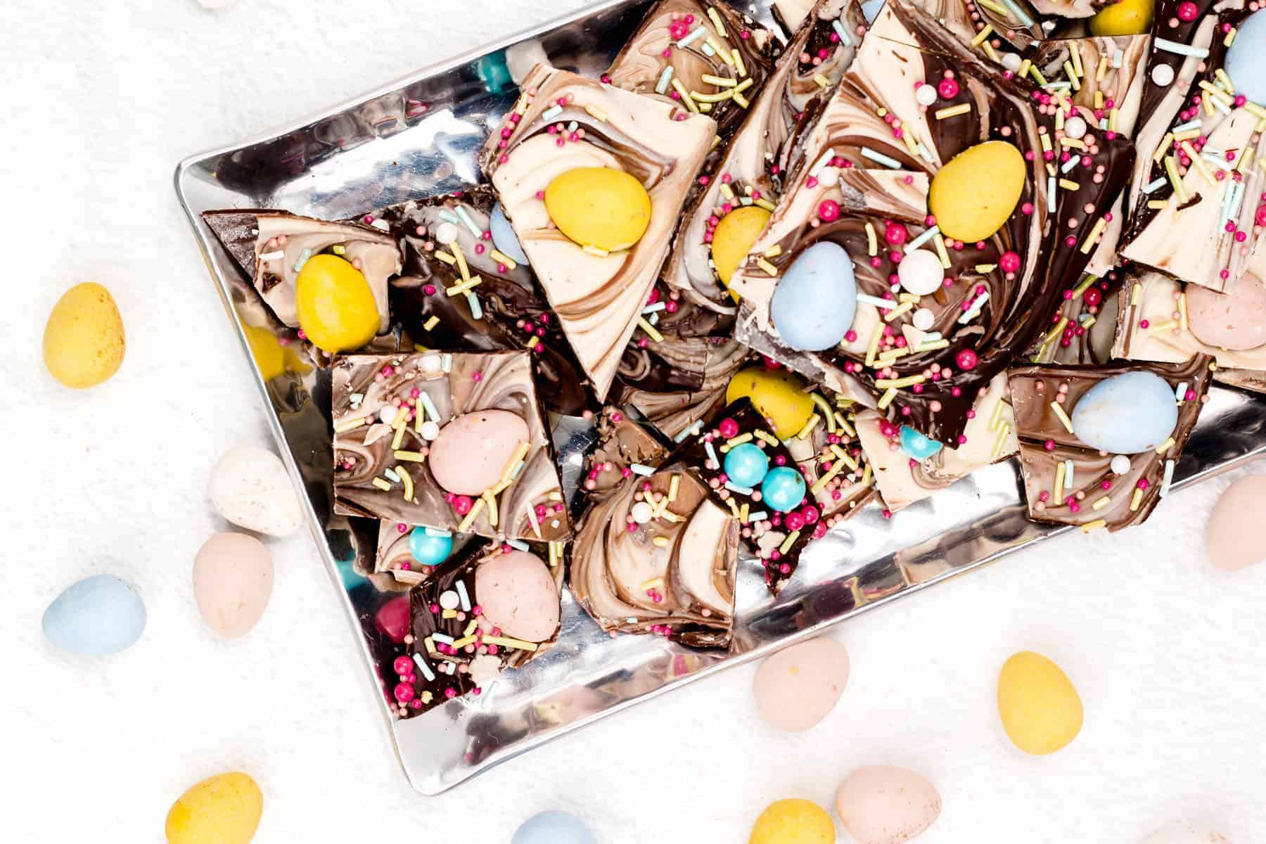 A rectangular tray filled with swirled chocolate bark pieces topped with colorful candy eggs and sprinkles, surrounded by scattered candy eggs on a white surface.