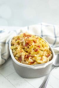 A ceramic dish filled with cooked cabbage and bacon pieces, placed on a white tiled surface with a spoon and a checkered cloth in the background.