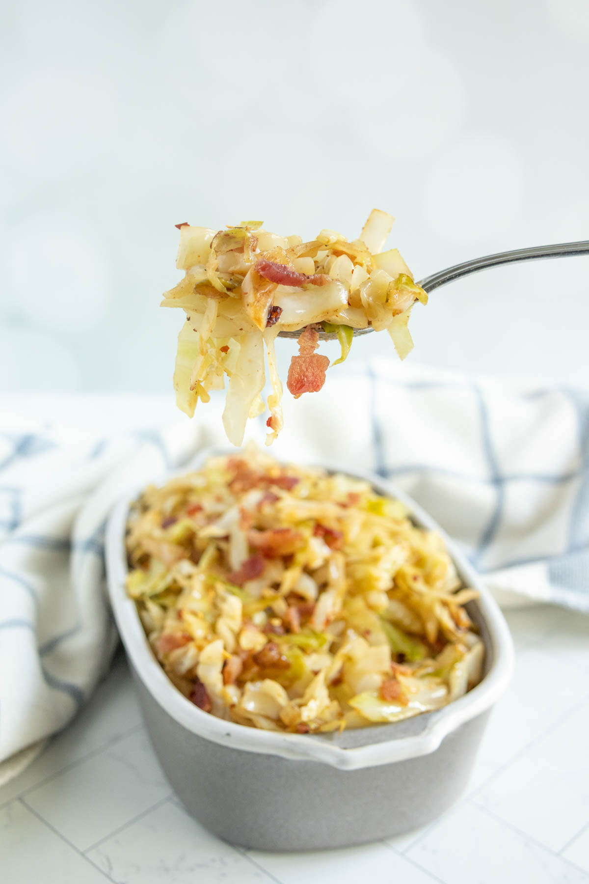 A fork holds a bite of cooked cabbage and bacon above a gray dish filled with the same cabbage and bacon mixture, with a white and blue cloth in the background.