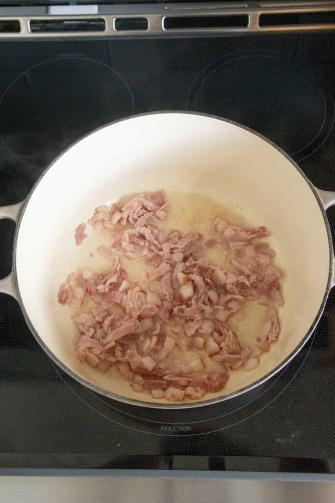 Chopped bacon pieces cooking in a white pot on a stove, starting to brown and release fat.