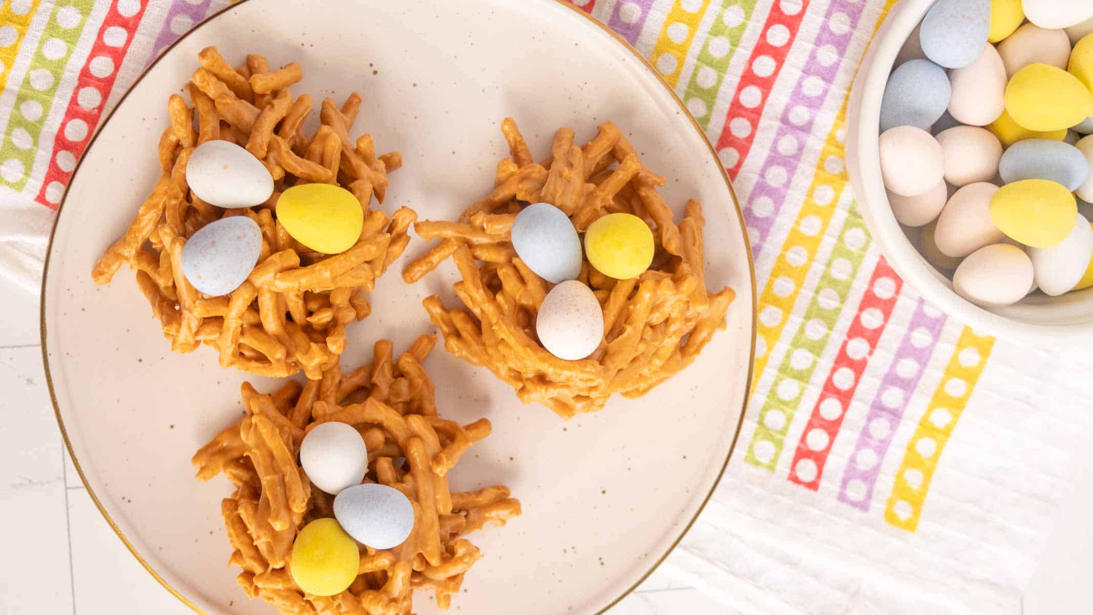 Three bird nest-shaped treats made of pretzel sticks and candy eggs on a plate, with a bowl of pastel-colored candy eggs nearby on a striped tablecloth.