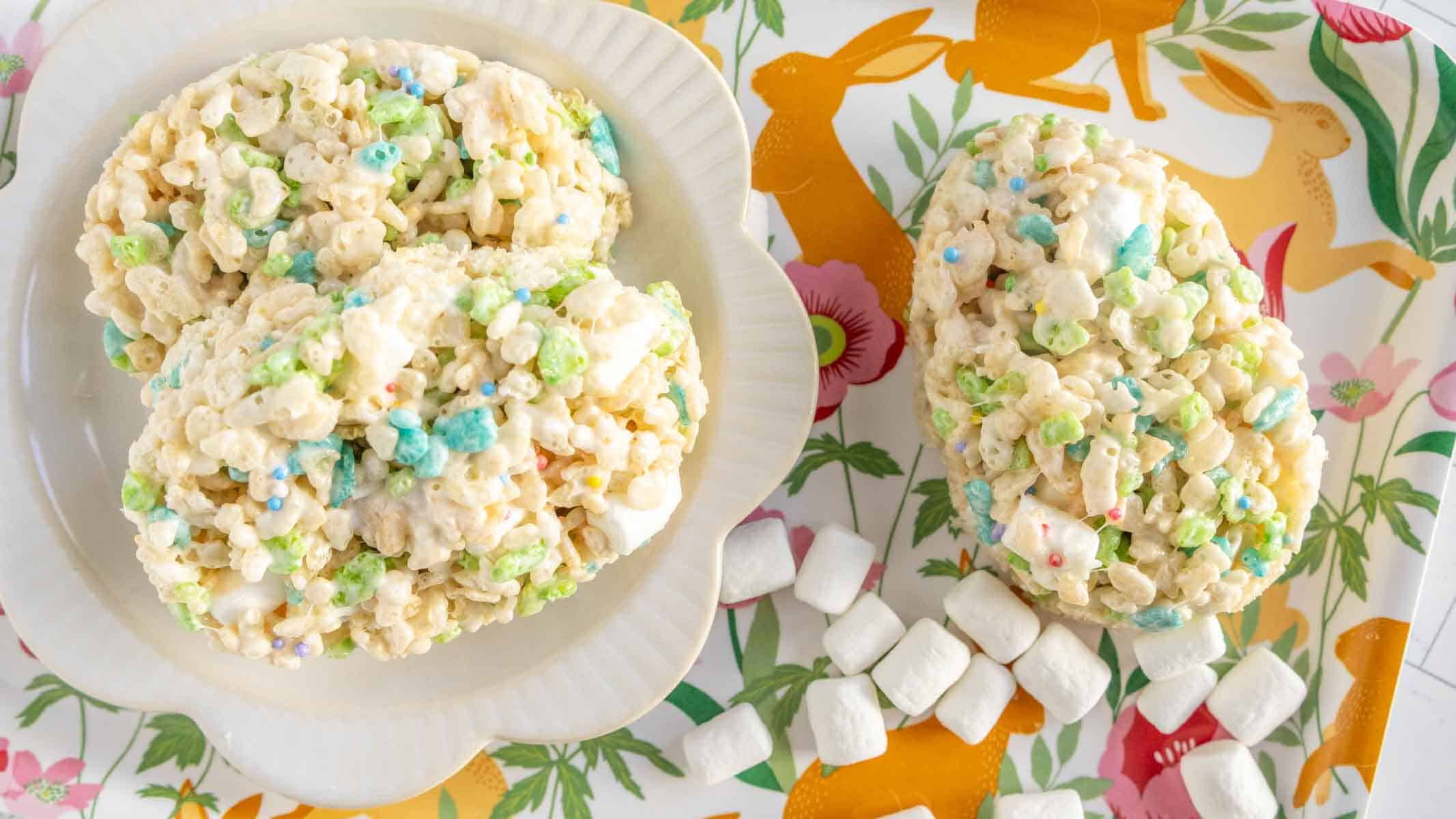 Two oval-shaped marshmallow cereal treats on a plate and one on a tray with scattered mini marshmallows, with a colorful spring-themed tray underneath.