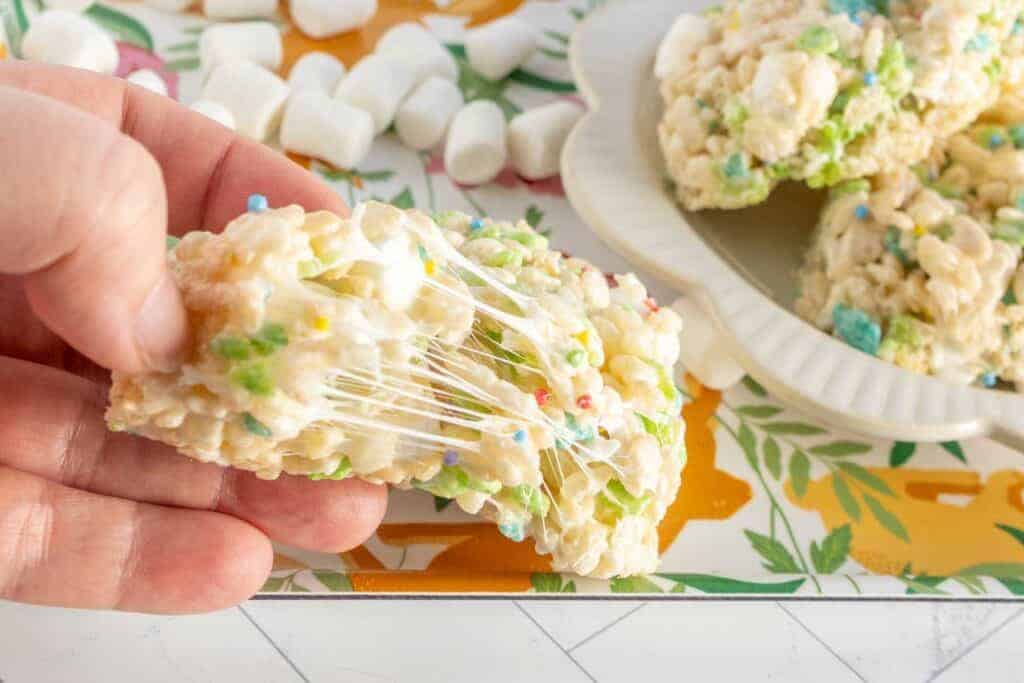 A hand pulls apart a gooey rice cereal treat with visible melted marshmallow; mini marshmallows and more Easter treats are in the background.