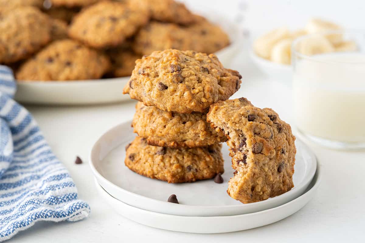 A stack of three oatmeal chocolate chip cookies on a plate, with one cookie partially eaten, next to a glass of milk and more cookies in the background.