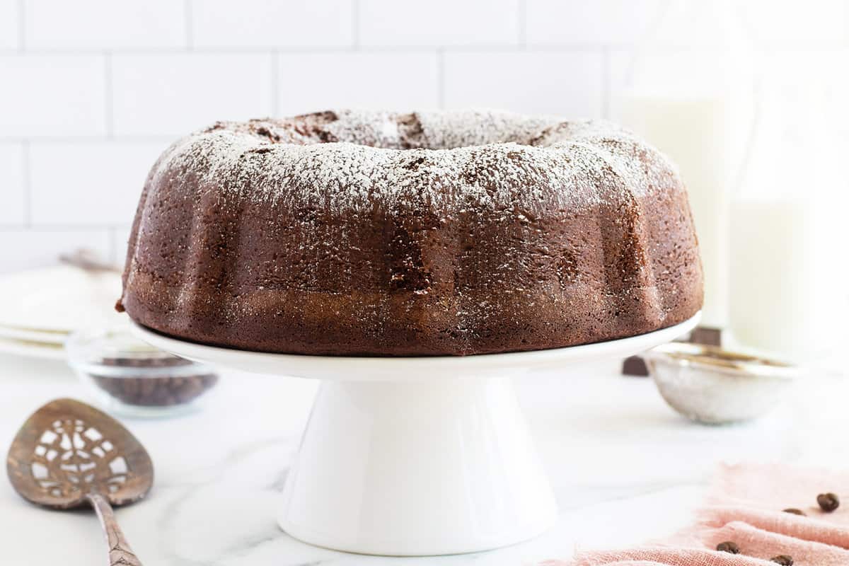 A chocolate Bundt cake dusted with powdered sugar sits on a white cake stand, with kitchen utensils and plates in the background.