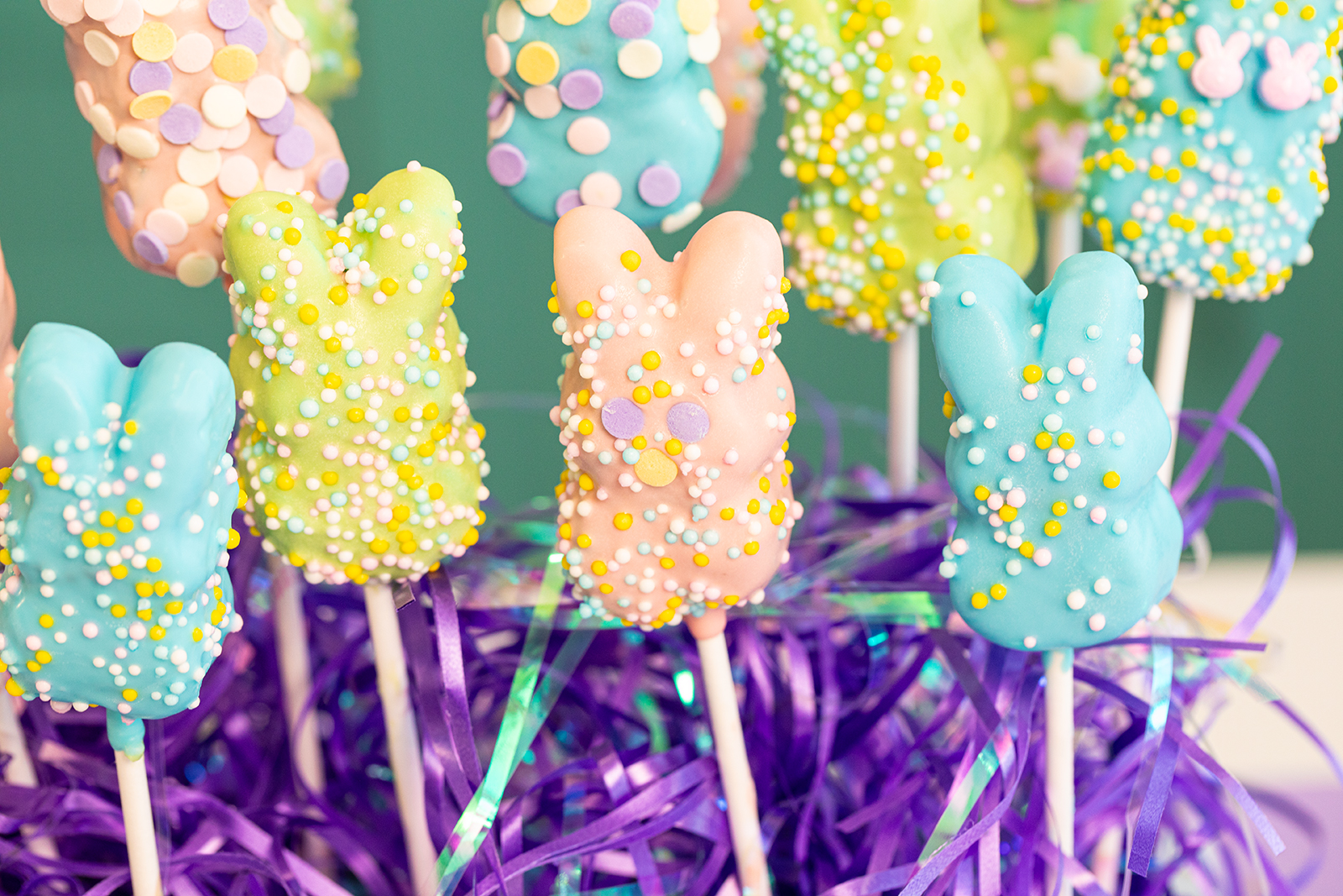 Colorful bunny-shaped treats on sticks, coated in pastel chocolate and sprinkles, are displayed upright in purple shredded paper.