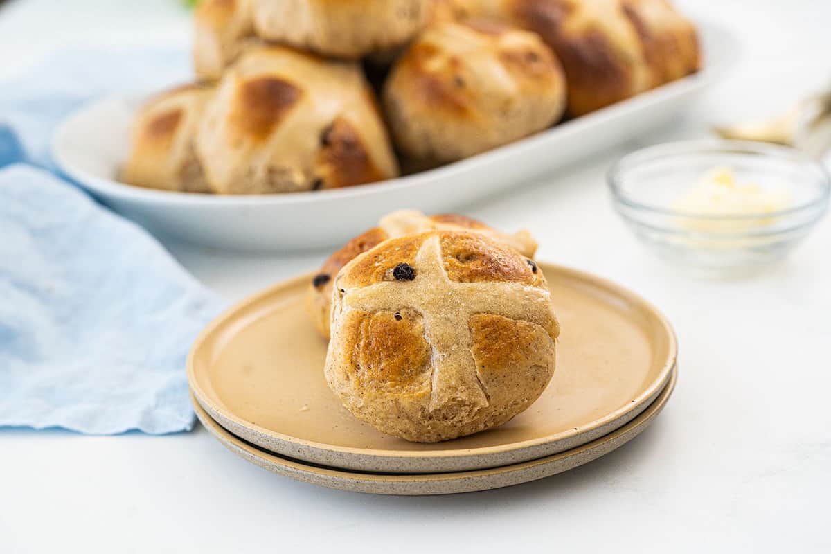 A close-up of two hot cross buns on stacked beige plates, with more buns on a white platter and a small dish of butter in the background.