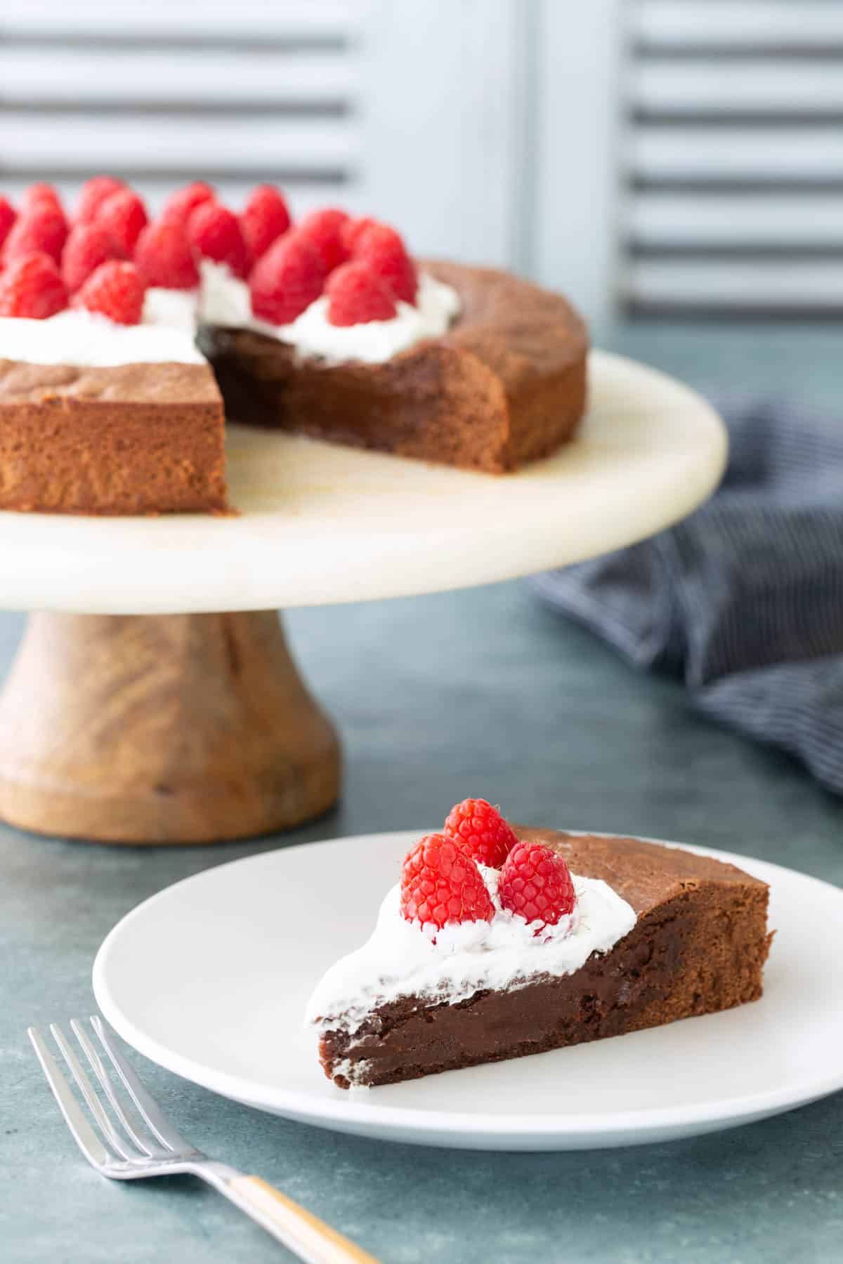 A slice of chocolate cake topped with whipped cream and raspberries is served on a plate, with the rest of the cake on a stand in the background.