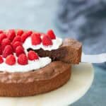 A slice of chocolate cake topped with whipped cream and fresh raspberries is being lifted from a cake stand.