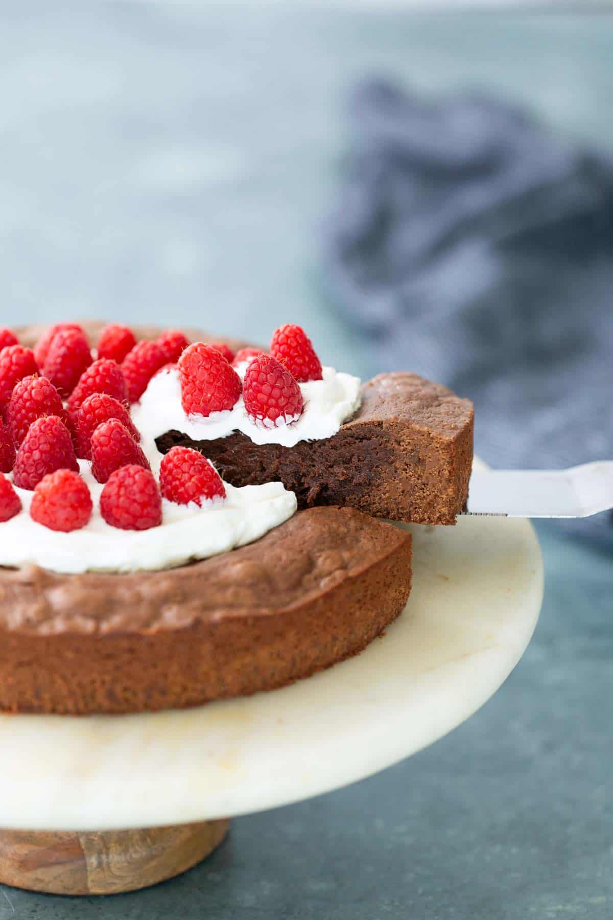 A slice of chocolate cake topped with whipped cream and fresh raspberries is being lifted from a cake stand.
