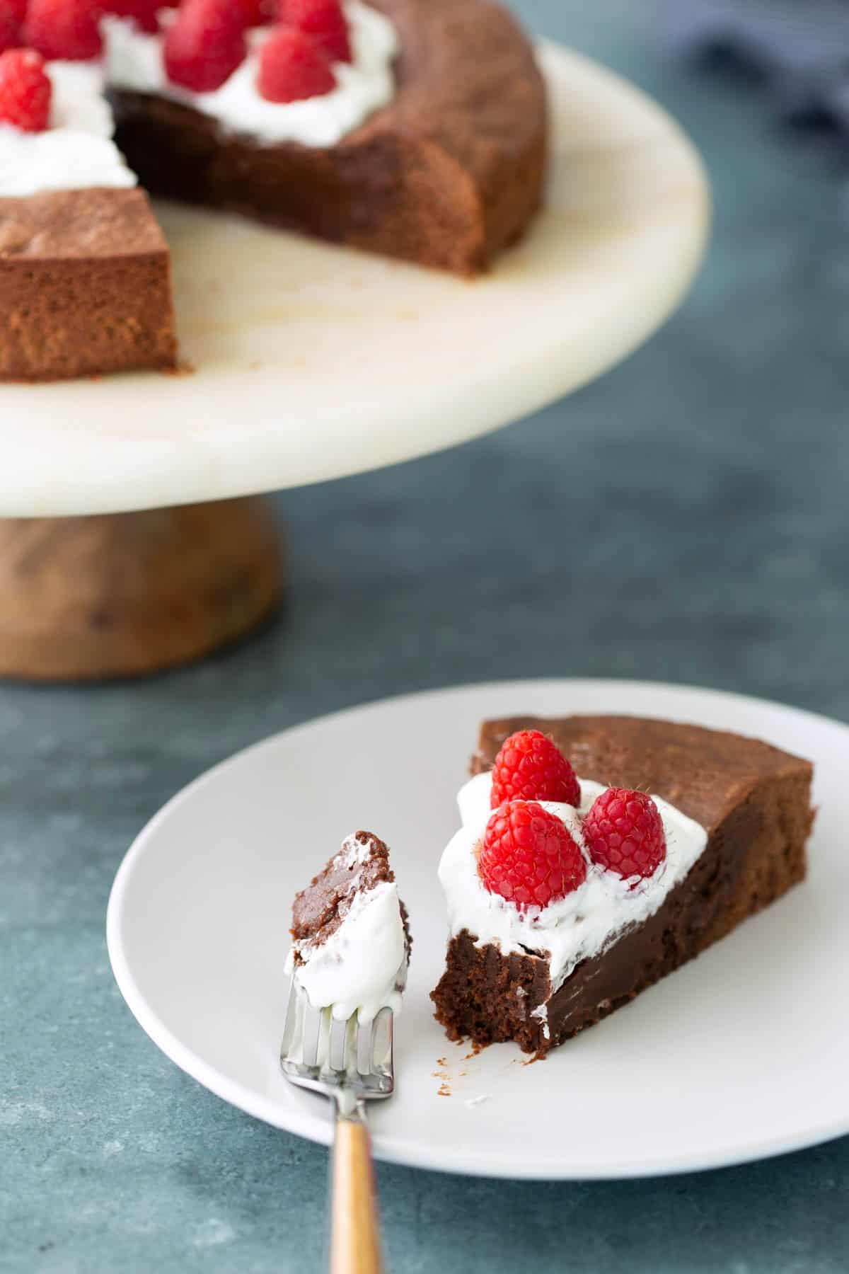 A slice of chocolate cake topped with whipped cream and raspberries sits on a white plate, with a fork holding a bite; the rest of the cake is on a stand in the background.