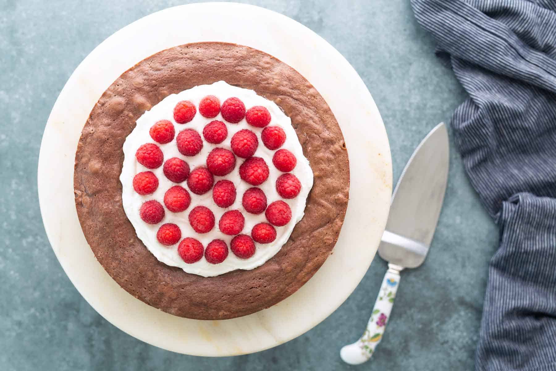 A chocolate cake topped with a circle of whipped cream and fresh raspberries sits on a marble platter next to a cake server and a striped cloth.