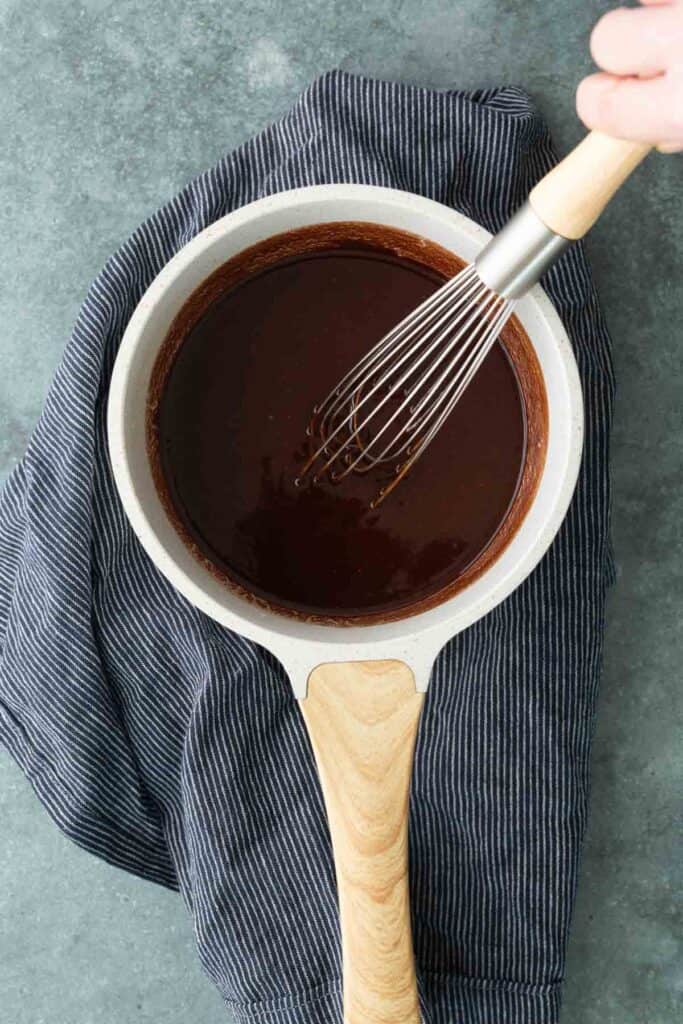 A hand whisking chocolate mixture in a saucepan placed on a striped cloth, viewed from above.