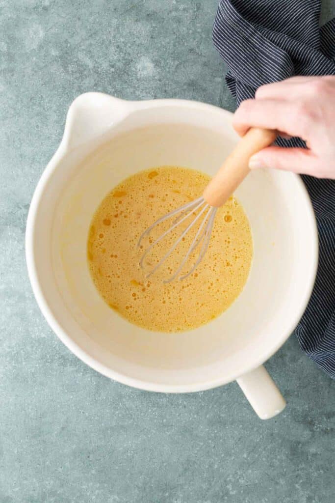 A hand holding a whisk mixes beaten eggs in a large white bowl on a blue surface with a striped cloth nearby.