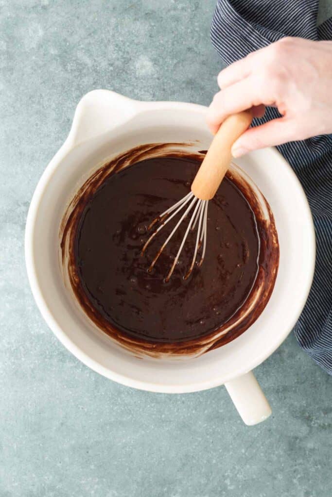 A hand holding a whisk mixes chocolate batter in a white mixing bowl on a gray countertop.