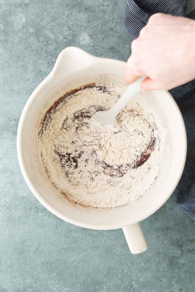 A hand stirs flour into a chocolate mixture in a white mixing bowl using a spatula, on a gray surface.