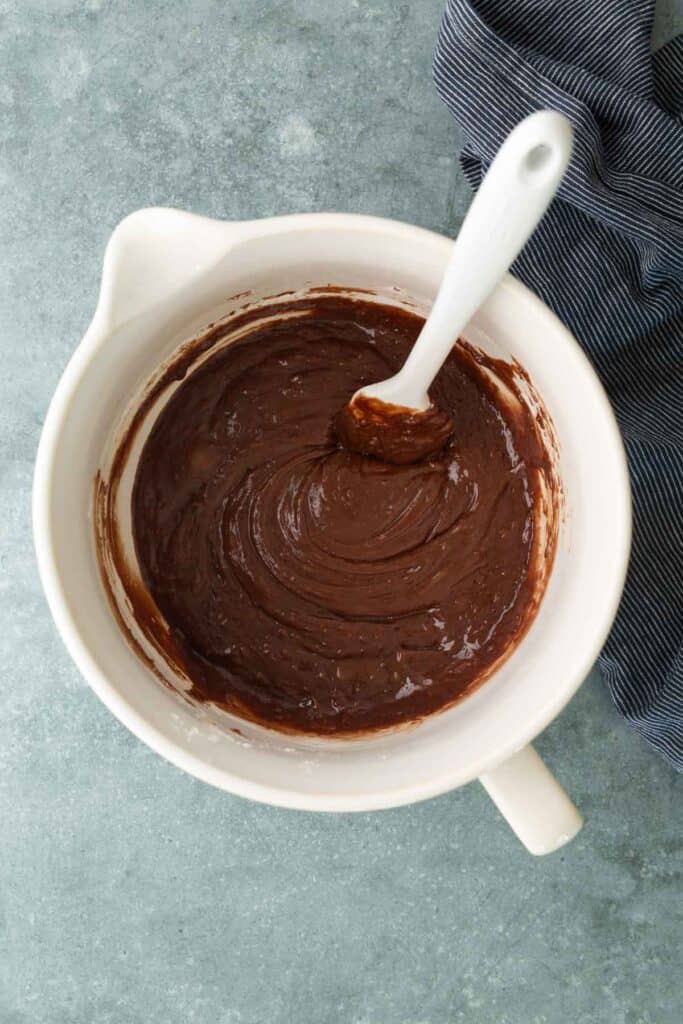 A white mixing bowl filled with chocolate batter and a white spatula, placed on a blue countertop next to a striped cloth.