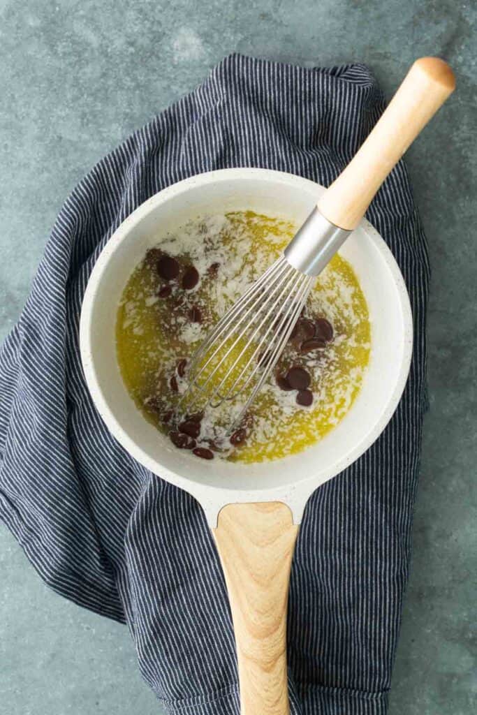 A whisk stirs chocolate chips into melted butter in a white saucepan, which sits on a striped cloth on a gray surface.