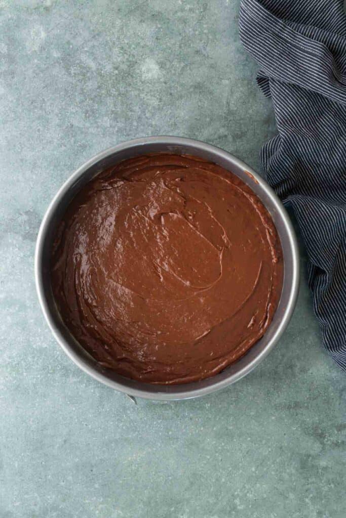 A round metal pan filled with smooth chocolate cake batter sits on a gray countertop next to a dark striped cloth.