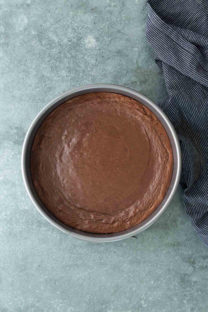 A round chocolate cake in a baking pan sits on a gray surface next to a dark striped cloth.