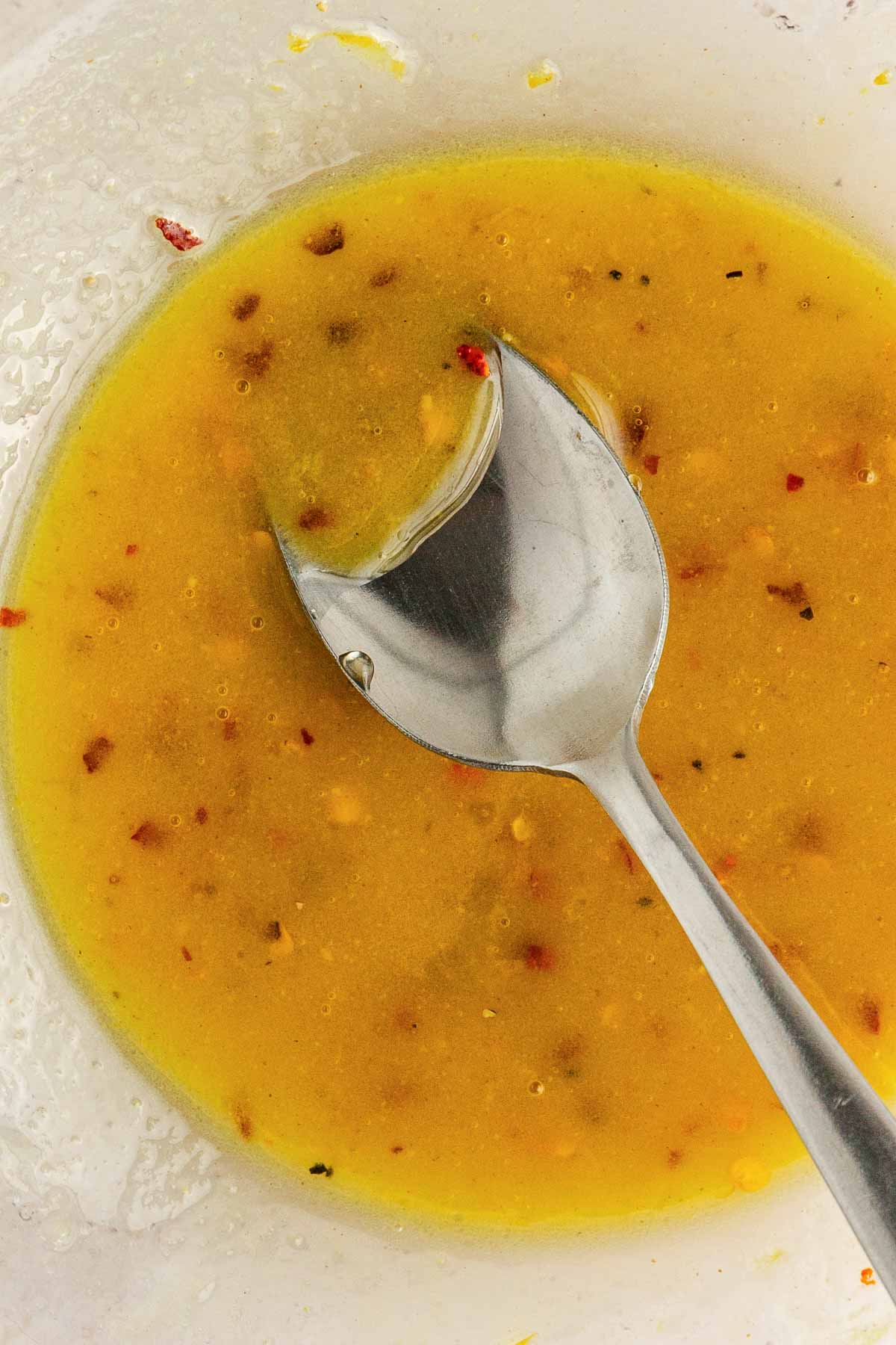 A metal spoon rests in a bowl of yellow vinaigrette with visible herbs and spices.