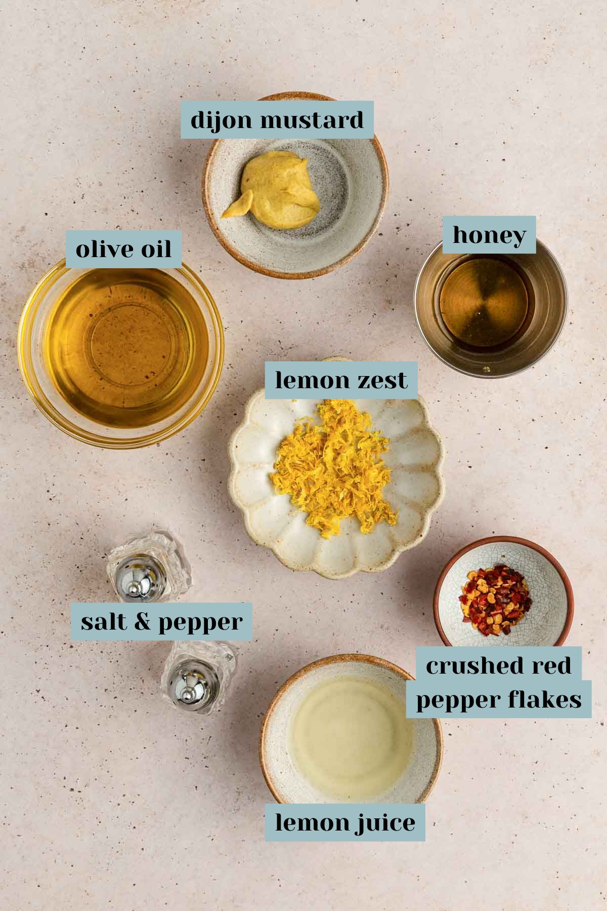 Top-down view of small bowls containing olive oil, Dijon mustard, honey, lemon zest, lemon juice, crushed red pepper flakes, salt, and pepper, each labeled.