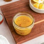 A small glass jar of yellow vinaigrette dressing sits on a wooden cutting board, with a dish of lemon wedges and a pepper grinder nearby.
