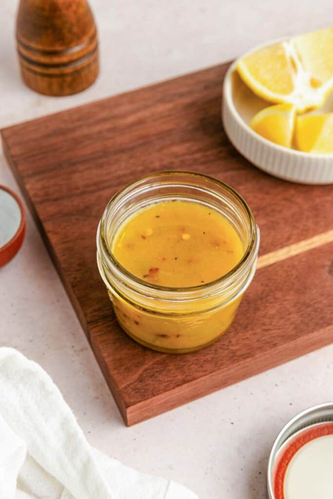 Lemon Salad Dressing A small glass jar of yellow vinaigrette dressing sits on a wooden cutting board, with a dish of lemon wedges and a pepper grinder nearby.