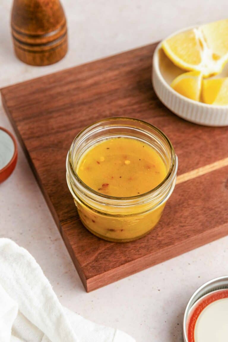 A small glass jar of yellow vinaigrette dressing sits on a wooden cutting board, with a dish of lemon wedges and a pepper grinder nearby.