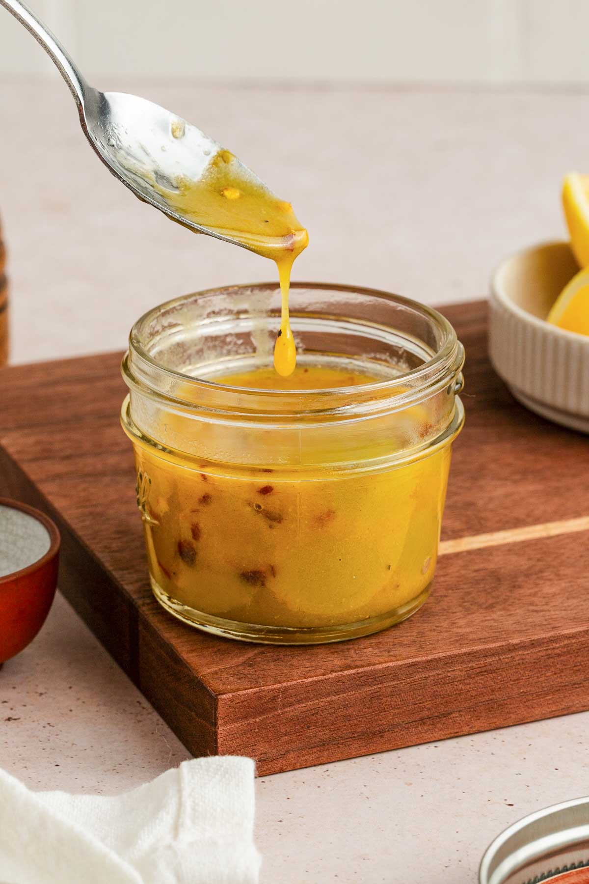 A spoon drizzling yellow vinaigrette dressing into a small glass jar on a wooden board, with lemon wedges in a bowl nearby.