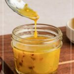 A spoon drizzling yellow lemon salad dressing into a small glass jar, sitting on a wooden surface next to a bowl.