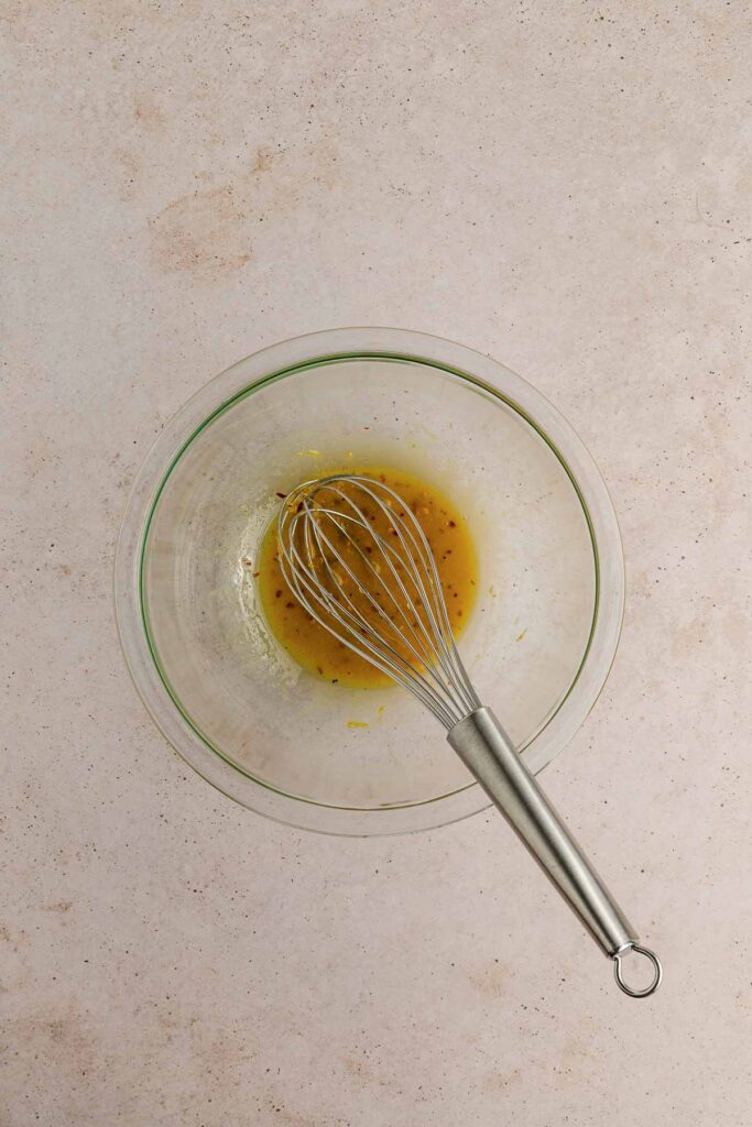 A glass bowl containing a small amount of light brown liquid with herbs, being whisked by a metal whisk, sits on a beige countertop.