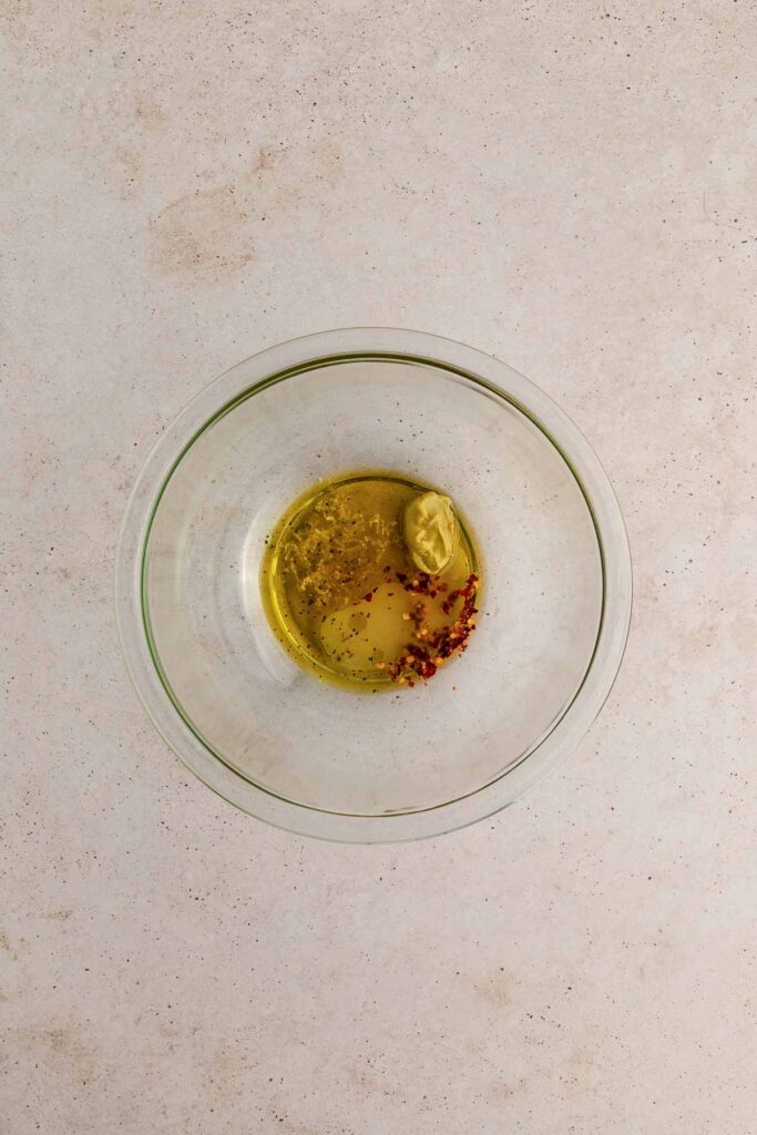 A glass bowl containing olive oil, mustard, red pepper flakes, and a light brown liquid on a light-colored countertop.