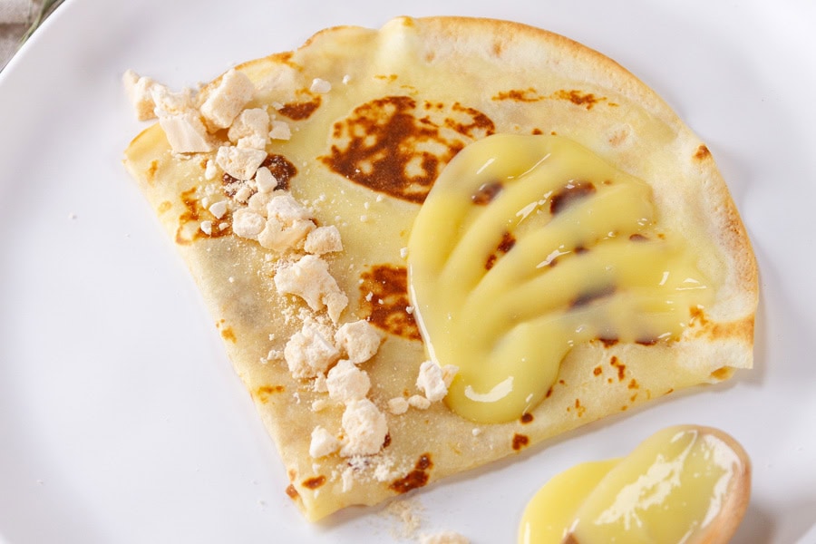 A folded crepe on a white plate topped with a yellow custard sauce and crushed meringue pieces.