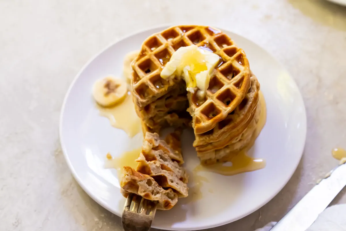 A stack of waffles topped with butter and syrup sits on a white plate with sliced bananas and a fork taking a bite.
