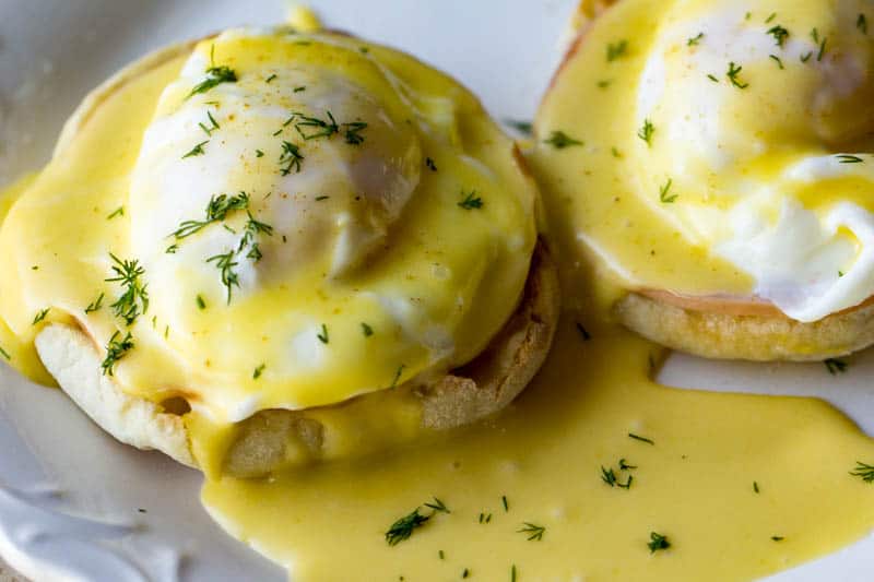 Two halves of an English muffin topped with poached eggs, ham, and yellow hollandaise sauce, garnished with chopped dill.